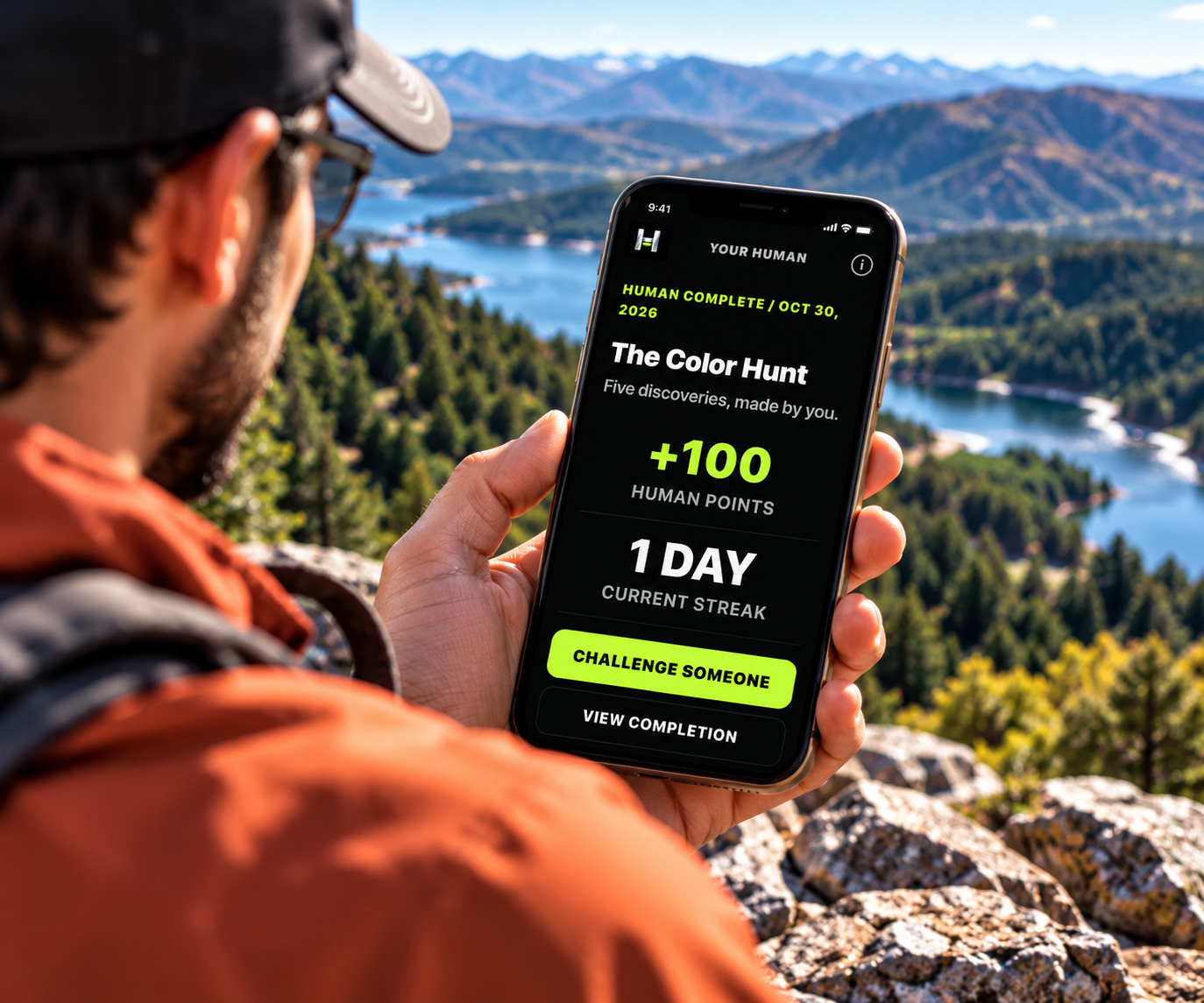 A hiker in an orange jacket sits at a mountain overlook above a lake, holding up a phone showing the HUMAN challenge he just finished