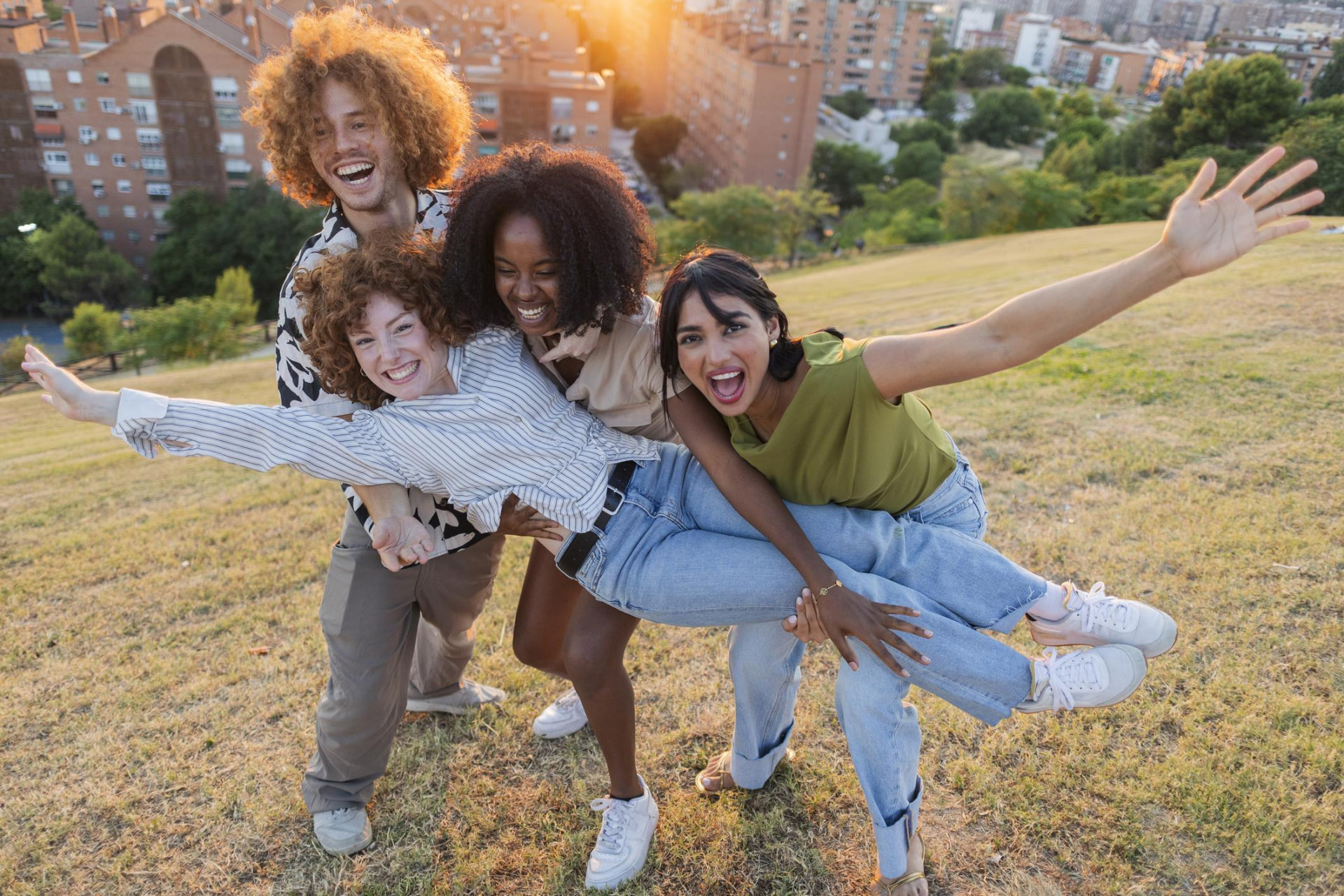 Four friends laugh and strike a flying pose together on a sunny hilltop above the city